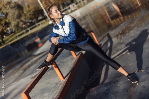 Fit slim woman in sportswear and headphones practicing leg stretching before training in a sports park