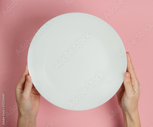 Female hand holds empty white plate on a pink pastel background. Top view.
