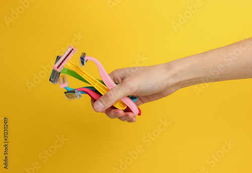 Female hand holds lot of colored disposable razors of epilators on yellow background. Beauty and fashion shot