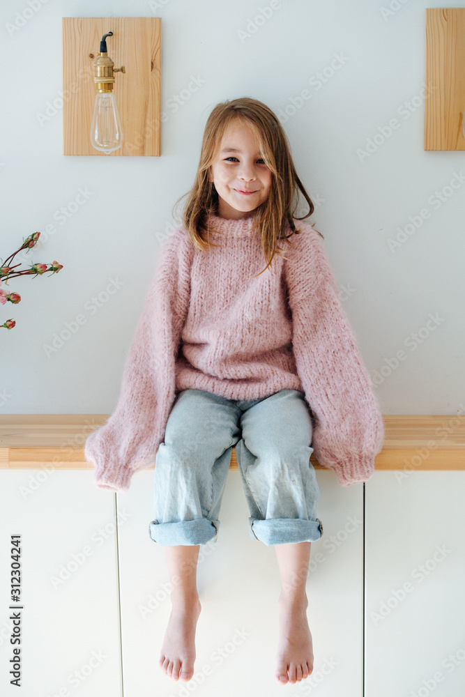 Happy smiling cute little barefoot girl in a pink oversized knitted sweater  sitting on a shelf