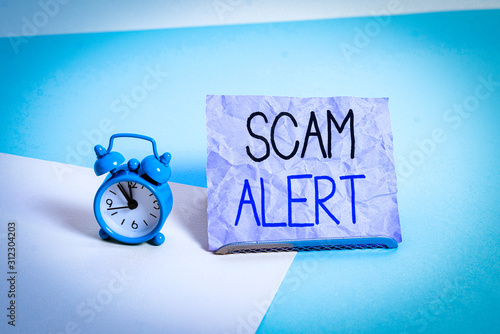 Handwriting text writing Scam Alert. Conceptual photo warning someone about scheme or fraud notice any unusual Mini size alarm clock beside a Paper sheet placed tilted on pastel backdrop photo