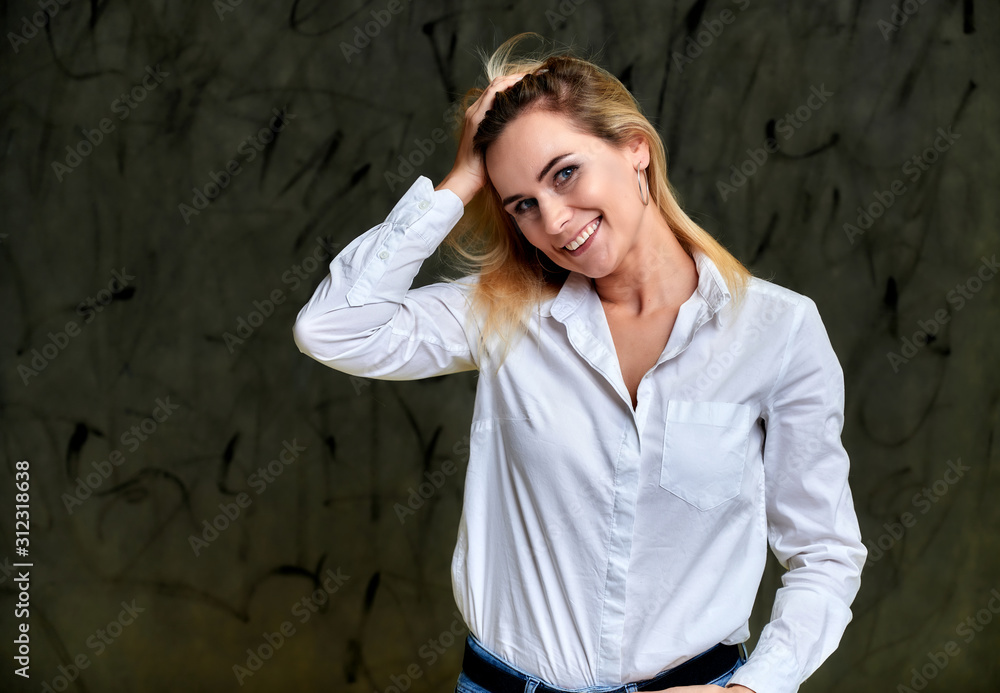 Portrait of a pretty smiling blonde woman in a white shirt with flying hair standing on a gray alternative background. Right in front of the camera with emotions. Portrait of a pretty smiling blonde woman in a white shirt with flying hair standing on a gray alternative background. Right in front of the camera with emotions.
