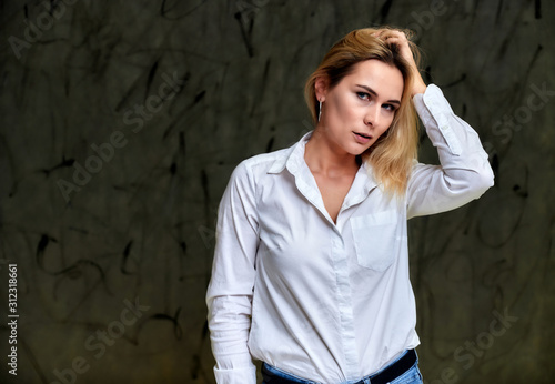Portrait of a pretty smiling blonde woman in a white shirt with flying hair standing on a gray alternative background. Right in front of the camera with emotions.