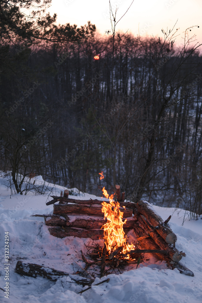 Campfire burns in the snow in the forest hill, on a background of snow covered trees and mountains campfire burning in cold winter. night fire and tent. Hiking Tourism. Flames, vertical, background