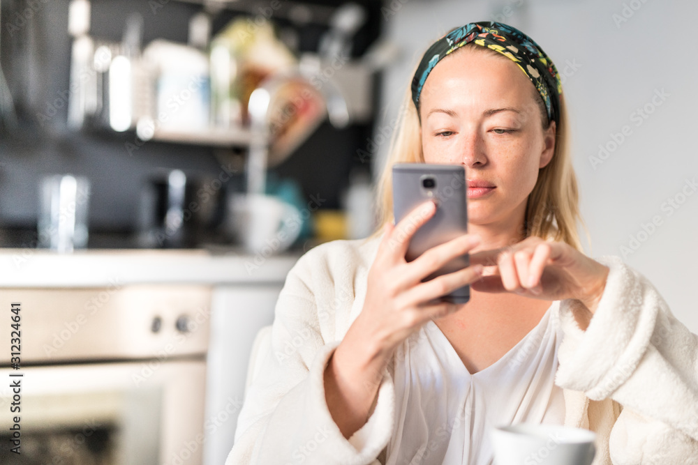 Beautiful caucasian woman at home, feeling comfortable wearing white bathrobe, taking some time to herself, drinking morning coffee and reading news on mobile phone device in the morning. - obrazy, fototapety, plakaty 
