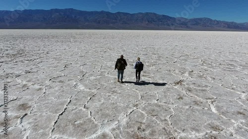 Young man and woman wanderlusts with touristic backpacks walking on dry surface of death walley and discussing view, hipster guys in wild environment of desolate salt basin Badwater explore USA nature photo