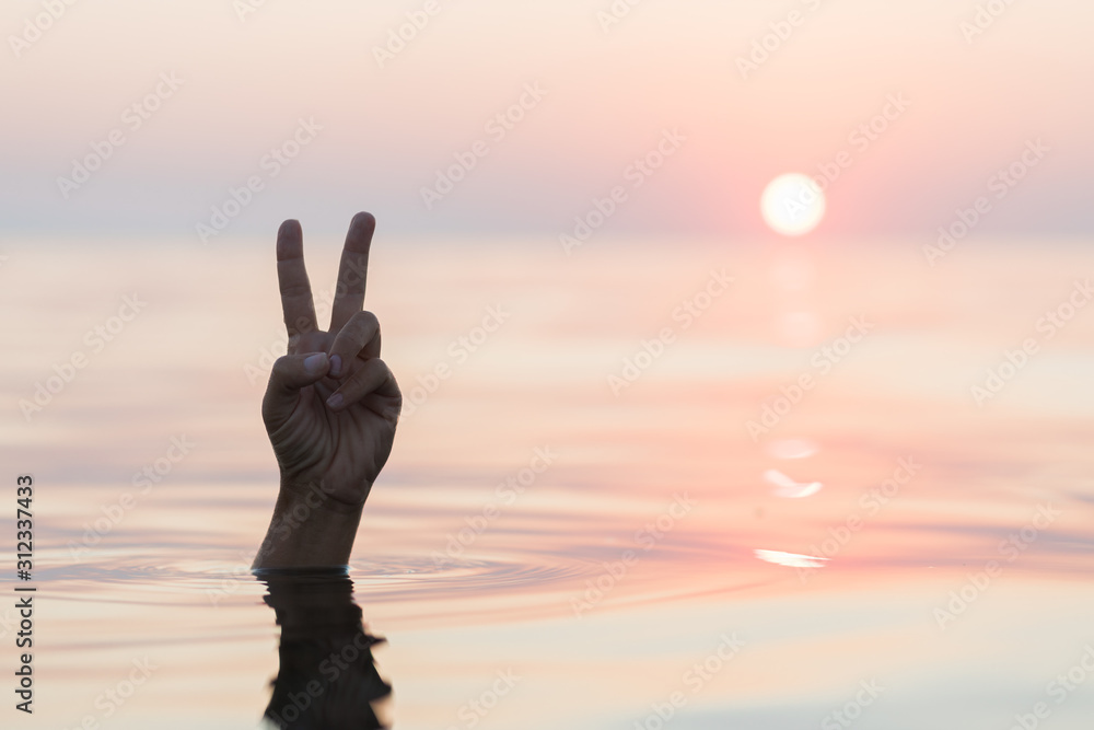 Hand emerging from the sea showing a peace sign in sunset
