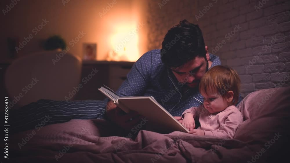 cheerful father is reading the bedtime stories to infant baby girl in bed at night cheerful father is reading the bedtime stories to infant baby girl in bed at night
