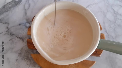 Frothing plant milk for morning latte coffee in an enamel saucepan with electric frother. Top view
