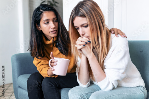 Slika na platnu Pretty young woman supporting and comforting her sad friend while sitting on the sofa at home