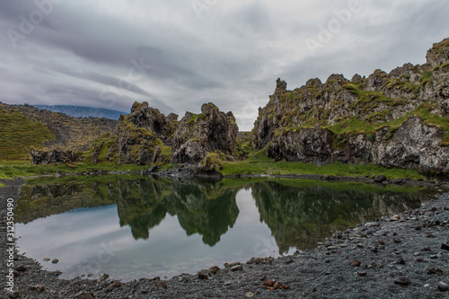 A small pond among volcanic stones in a circle. The stones are covered with green grass. Stones reflected in a pond
