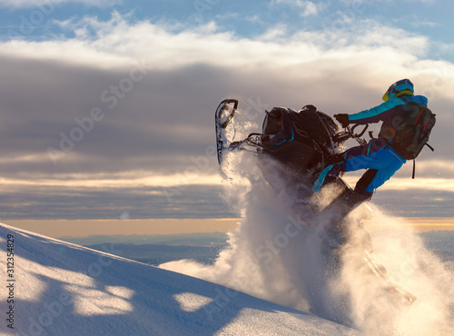 a snowmobile rider jumps in a mountain valley at dawn. sports snow bike with snow splashes and snow trail. bright snowmobile and suit without brands. snowmobilers sports riding. stock photo