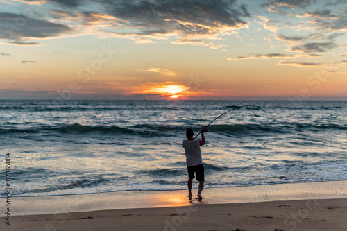 Fisherman stands on sandy beach during sunset