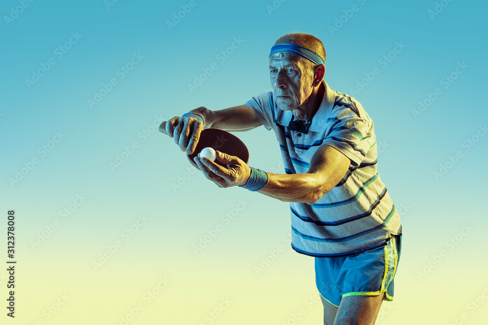 Fototapeta Senior man wearing sportwear playing table tennis on gradient background, neon light. Caucasian male model in great shape stays active. Concept of sport, activity, movement, wellbeing, confidence.