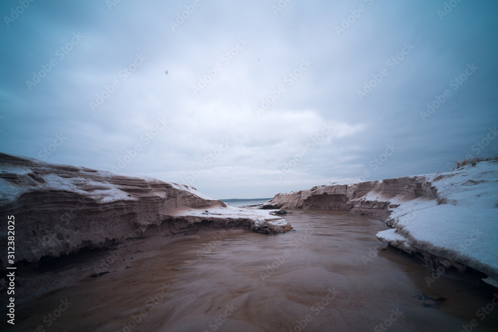 Tiny creek falls into Finnish Gulf though frozen sand dunes on Salmistu beach in Estonia. Empty coast in chilly winter morning, blue skyline over rural Nordic landscape and green conifer forest. Tiny creek falls into Finnish Gulf though frozen sand dunes on Salmistu beach in Estonia. Empty coast in chilly winter morning, blue skyline over rural Nordic landscape and green conifer forest.