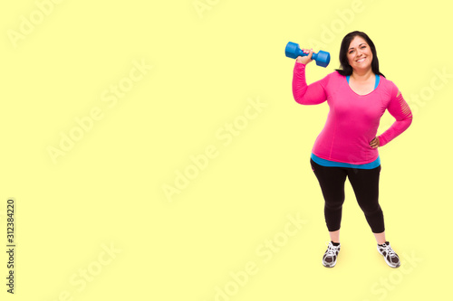 Middle Aged Hispanic Woman In Workout Clothes Holding Dumbbell Against A Bright Yellow Background