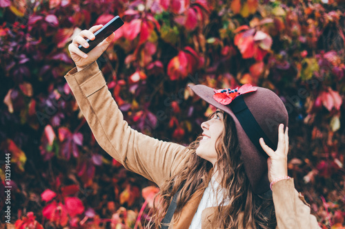 Woman in hat makes selfie on the background of autumn leaves.