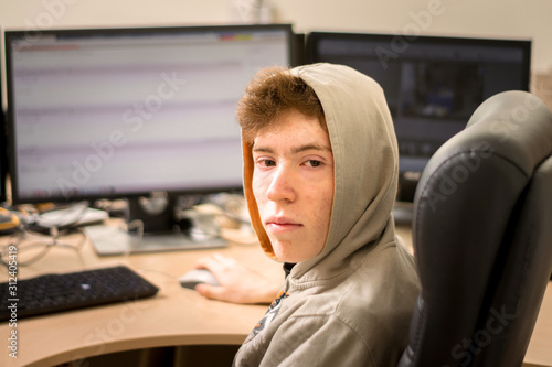 A portrait of a young guy in a hood is in front of the monitors. A teenager with facial pricks sits at a table with several displays. The guy sits in an office chair and works at the computer.
