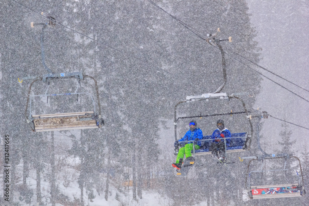 Winter landscape - view of the chairlift lift with skiers in the winter mountain forest during a snowfall