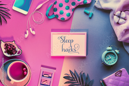 Healthy night sleep creative concept. Sleeping mask, alarm clock, earphones, earplugs, calming tea and pills. Split two tone pink and green paper background with leaves. Text "Sleep hacks" in frame.