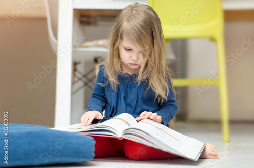 Little fair-haired girl is reading a book while sitting on the floor at home. Early childhood education