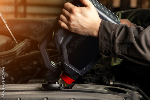 A male worker pours motor oil into a car engine from a gray canister with a red cap under the hood during maintenance and repair. Auto service industry.