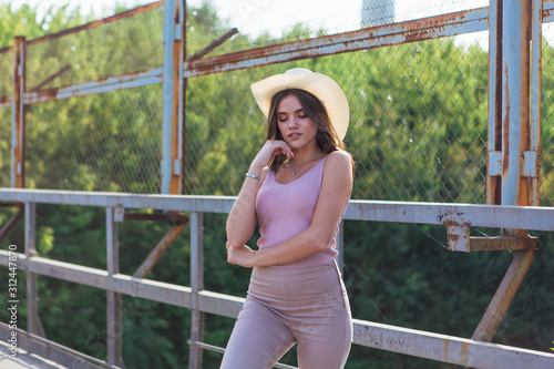 Pretty young woman wearing cowboy hat posing on the old rusty bridge during sunset.