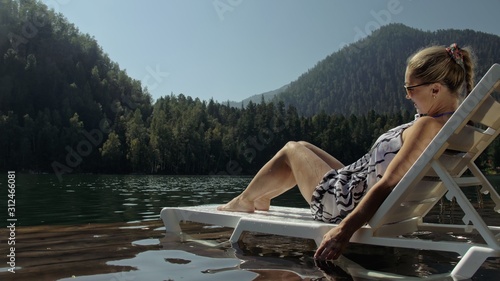 Woman lie on a sunbed in sunglasses and a boho silk shawl. Girl rest on a flood wood underwater pier. The pavement is covered with water in the lake. In the background are mountain and a forest.