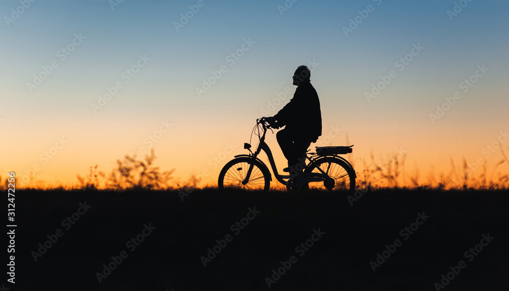 Obraz premium Male cyclist on the e-bike or electric bicycle on the sunset background riding up the hill. Silhouette of the old man in profile. Active pension. Travel. Sport. Copy space.