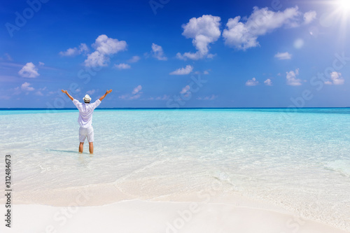 A happy man in white summer clothes stands with outstretched arms in turquoise  tropical waters in the Maldives islands  Indian Ocean