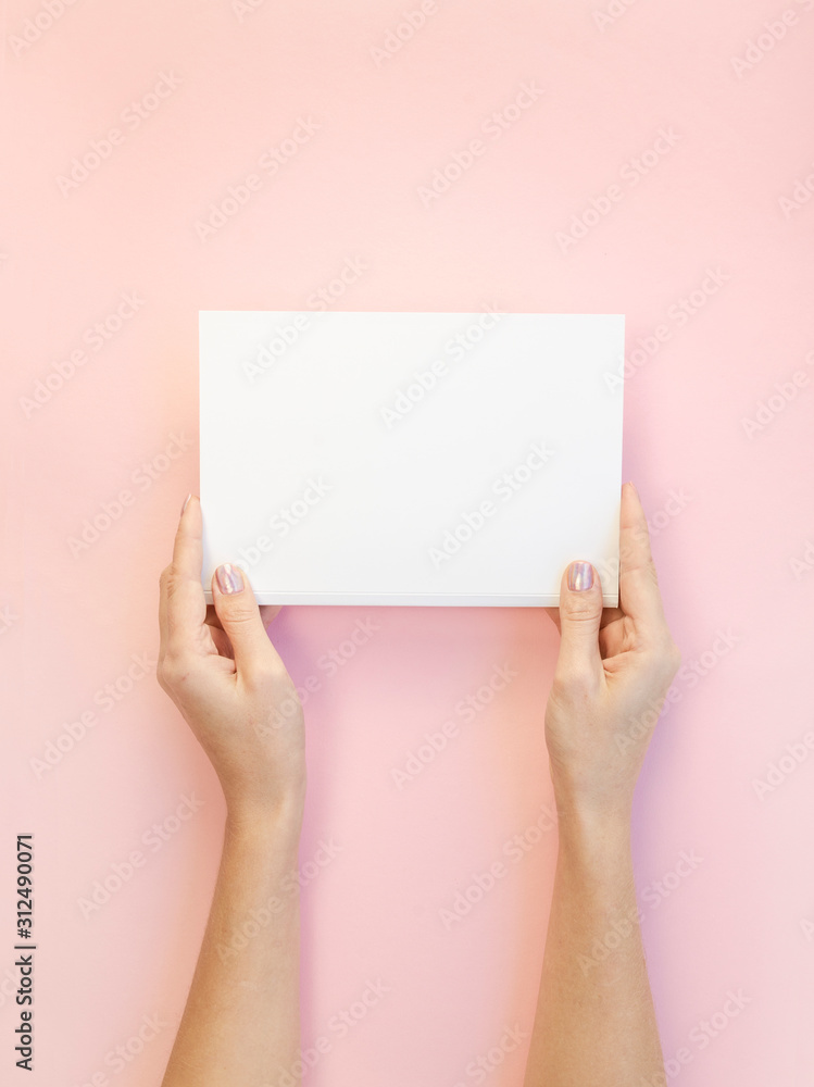 Vertical mockup female hands hold white blank paper or magazine on a pastel pink background. Valentine's day card or gift concept with copy space Vertical mockup female hands hold white blank paper or magazine on a pastel pink background. Valentine's day card or gift concept with copy space