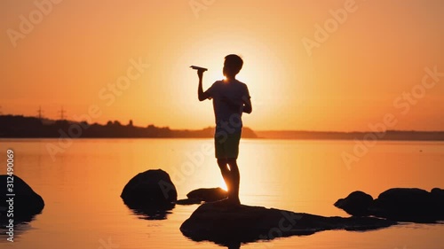 Silhouette of a boy with a paper plane at sunset. Child is standing on a stone on the orange river background. Bright rays of evening sun shines on a boy.