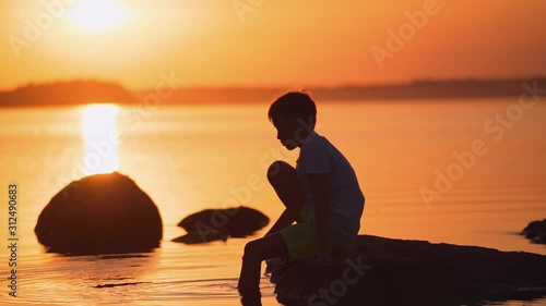 Little boy sitting on a stone among the water. Silhouette of a child near the river at a beautiful orange sunset. Slow motion.