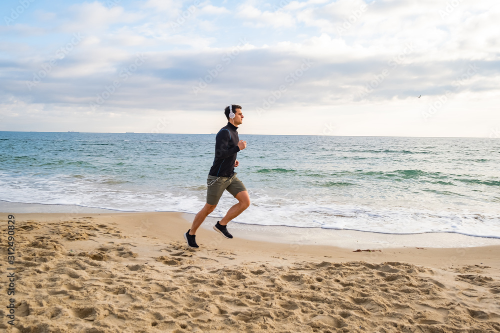 Fit male runner training on the summer beach and listen to music against beautidul sky and sea Fit male runner training on the summer beach and listen to music against beautidul sky and sea