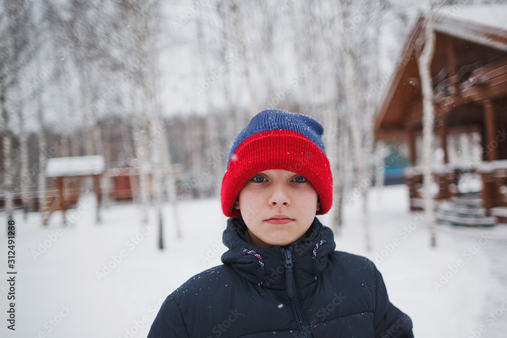A sad young happy teenage boy in a blue winter jacket and a colored hat in winter outdoors. Frost winter season.