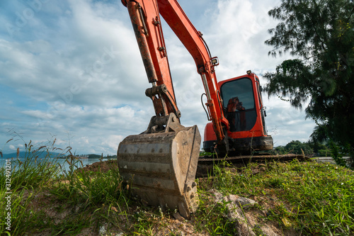 Small orange excavator on a ground against blue sky and sea for a works on construction site. Small tracked excavator standing on a ground with a blue sea on background. Heavy industry.