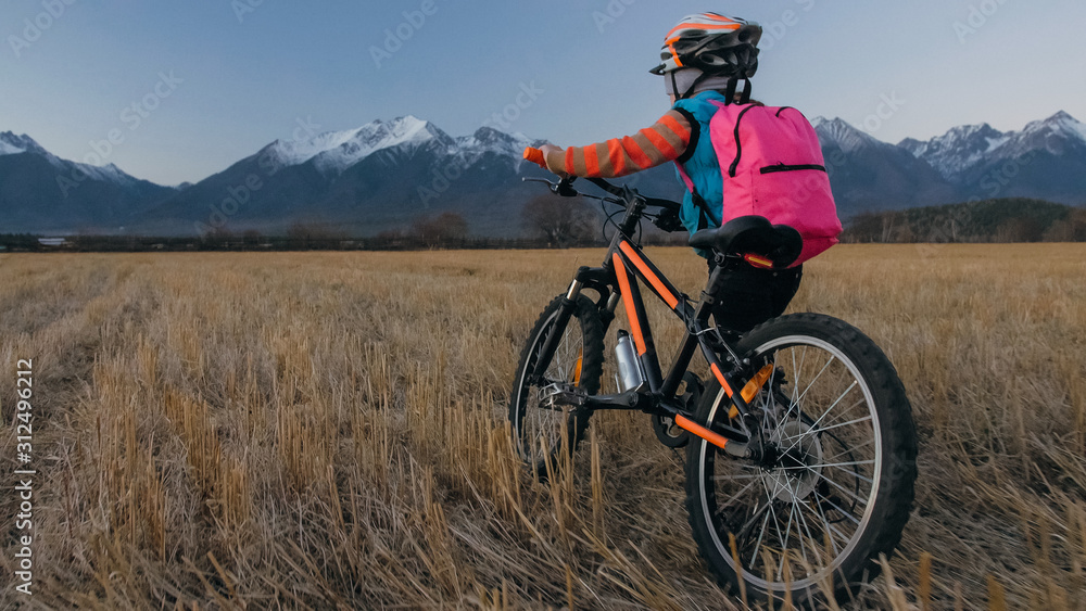 Obraz premium One caucasian children walk with bike in wheat field. Little girl walking black orange cycle on background of beautiful snowy mountains. Biker stand with backpack and helmet. Mountain bike hardtail.