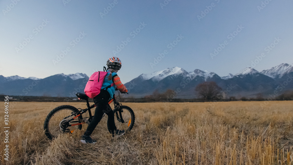 One caucasian children walk with bike in wheat field. Little girl walking black orange cycle on background of beautiful snowy mountains. Biker stand with backpack and helmet. Mountain bike hardtail.