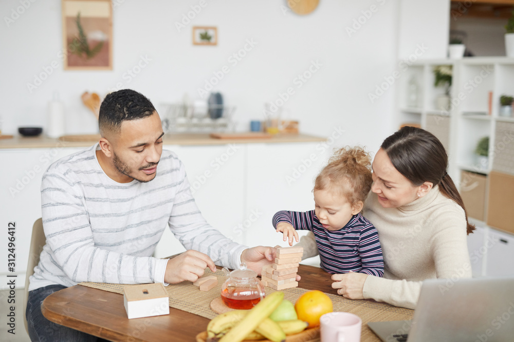 Portrait of happy mixed-race family playing with cute little daughter while enjoying breakfast at home, copy space