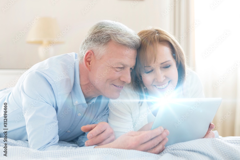 Obraz premium Smiling mature couple sharing digital tablet while lying on bed at home