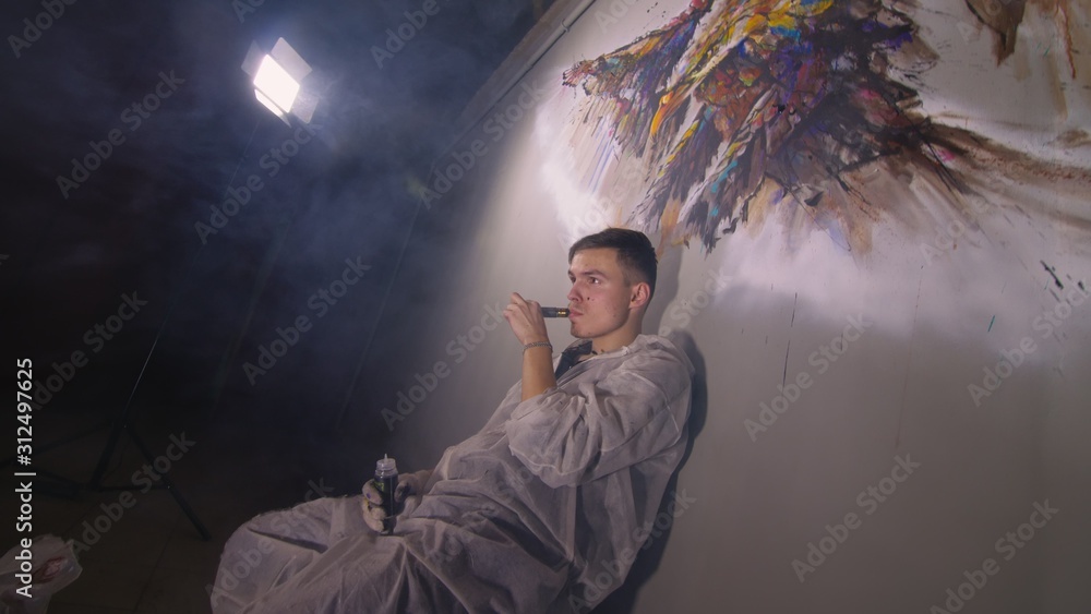 Artist designer draws an eagle on the wall. Craftsman decorator paints a picture with acrylic oil color. Vaper smokes vape. Painter painter dressed in a paint coat. Indoor. Dark magic cinematic look. Artist designer draws an eagle on the wall. Craftsman decorator paints a picture with acrylic oil color. Vaper smokes vape. Painter painter dressed in a paint coat. Indoor. Dark magic cinematic look.