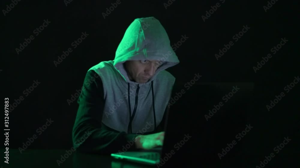 Man is a guy, a hacker in the hood, at the computer, looks at the monitor and prints codes. Man is a guy, a hacker in the hood, at the computer, looks at the monitor and prints codes.