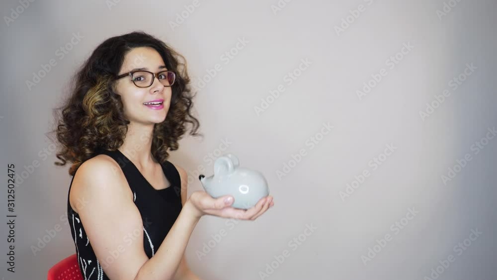 Young cute girl with dark curly hair holding mouse shaped piggy bank, shows gesture ok and points a finger at empty space. Girl holding a piggy bank with coins and shows ok. Copy space 4k Young cute girl with dark curly hair holding mouse shaped piggy bank, shows gesture ok and points a finger at empty space. Girl holding a piggy bank with coins and shows ok. Copy space 4k