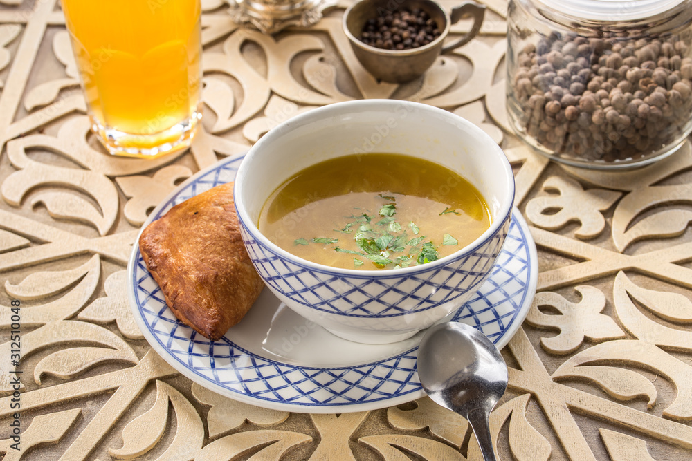 chicken broth with bread bun and parsley in bowl with glass of yellow drink on oriental wooden table chicken broth with bread bun and parsley in bowl with glass of yellow drink on oriental wooden table