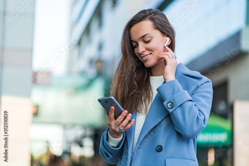 Stylish trendy happy joyful brunette woman in a blue coat holds a smartphone and using wireless white headphones for listen music in the city centre. Modern people