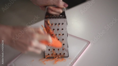 Hands of a young girl rub an orange carrot using a grater. Preparing fresh carrots before cooking on kitchen.