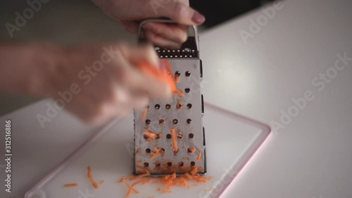 Hands of a young girl rub an orange carrot using a grater. Preparing fresh carrots before cooking on kitchen.