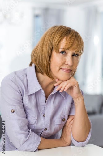 Obraz na plátně Portrait of mature woman with hand on chin sitting at table in house