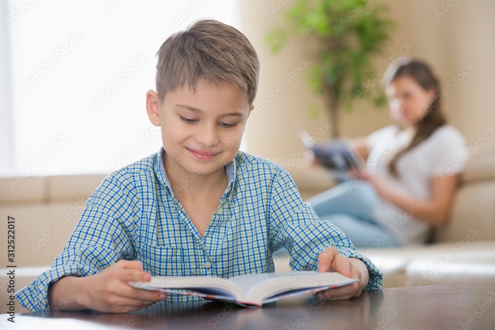 Cute boy reading book with mother in background at home Cute boy reading book with mother in background at home