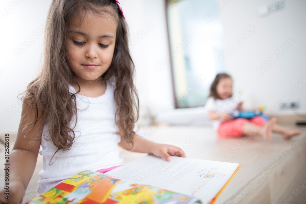 Cute girl reading book with sister in background at home Cute girl reading book with sister in background at home